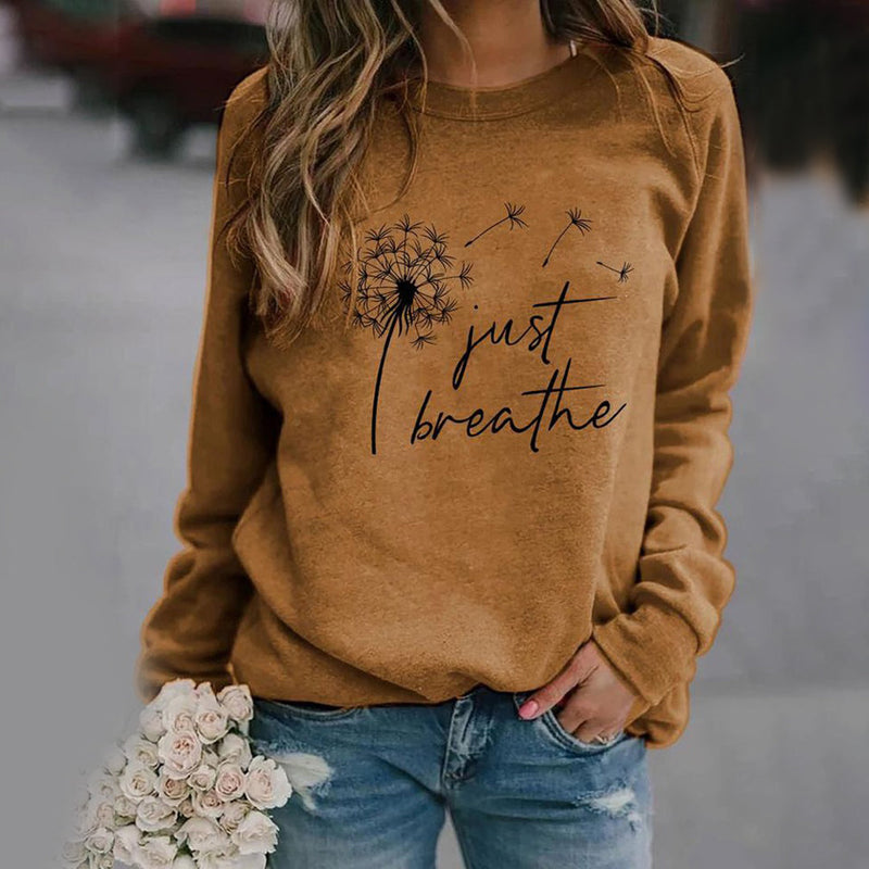 Frau trägt braunes Sweatshirt mit Löwenzahn-Design und "Just Breathe"-Aufdruck, lässige Mode.