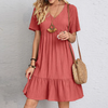 Frau in rotem Sommerkleid mit V-Ausschnitt, Strohtasche und Halskette vor grauer Wand.