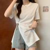 Frau in weißem T-Shirt mit Knoten-Detail und blauen Jeansshorts vor Holzwand.