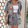 Frau in gemustertem Sommer-Zweiteiler, blau-orange, mit weißem Top, hält Strohtasche.